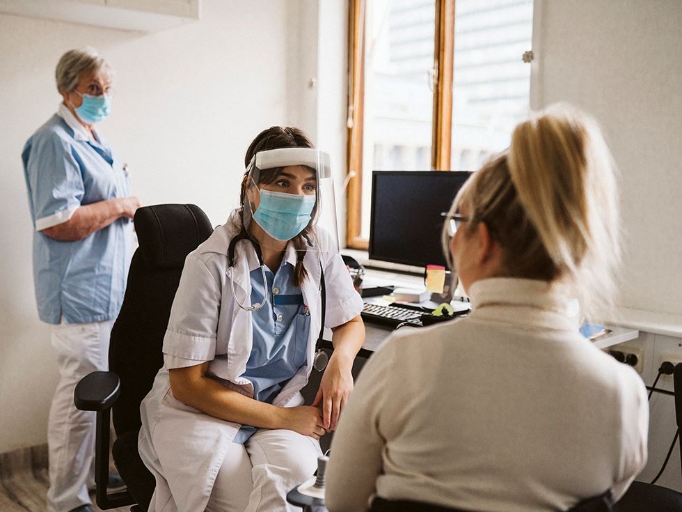 Kvinnlig läkare med munskydd pratar med patient på vårdcentral.