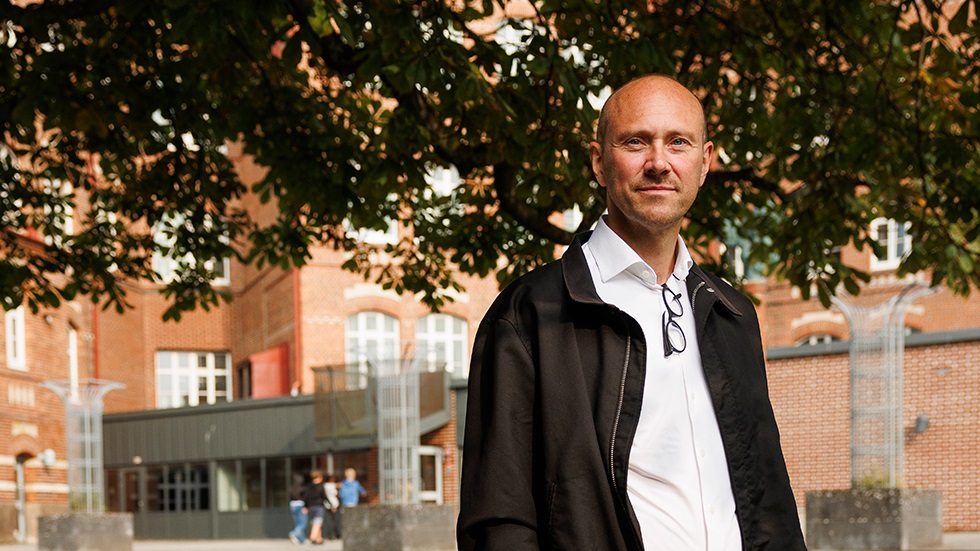 Porträtt på man i vit skjorta och mörk jacka utanför skolbyggnad. 