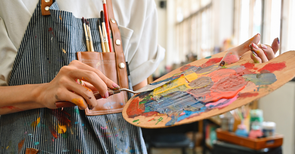 Foto av en person med ett kladdigt förkläde med penslar i bröstfickan. Personen håller i en palett med en massa färger.som håller i en palett med en massa färg