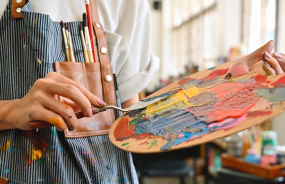 Foto av en person med ett kladdigt förkläde med penslar i bröstfickan. Personen håller i en palett med en massa färger.som håller i en palett med en massa färg