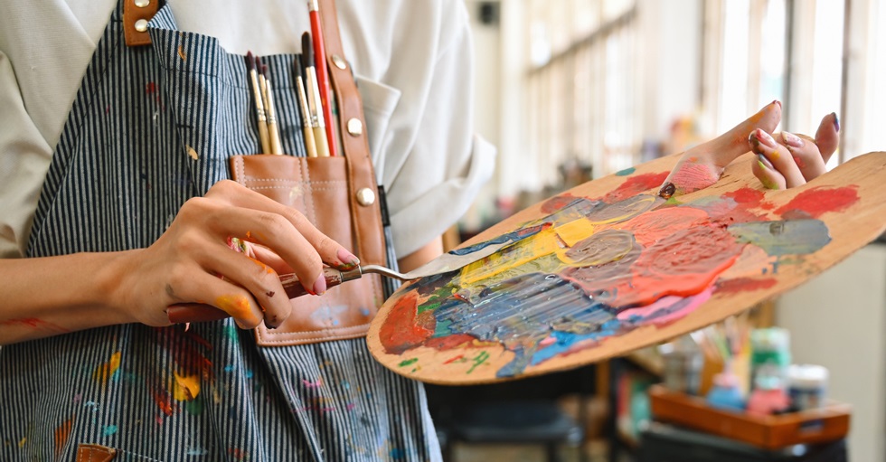 Foto av en person med ett kladdigt förkläde med penslar i bröstfickan. Personen håller i en palett med en massa färger.som håller i en palett med en massa färg