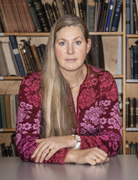 Porträtt av Cecilia Österman som sitter vid ett bord med en bokhylla bakom sig.