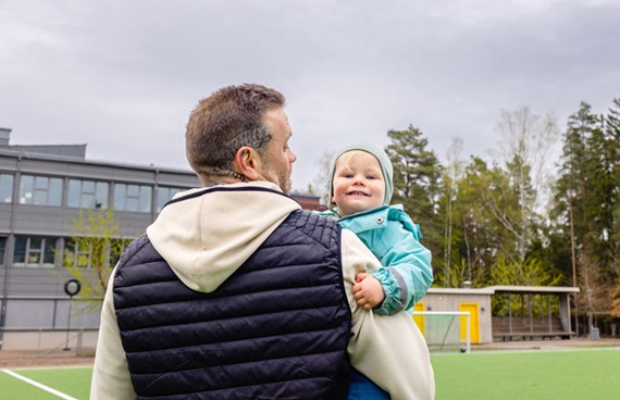 En pappa bakifrån med ett barn på armen som tittar in i kameran. Bilden är tagen på hösten på en fotbollsplan framför en skola.