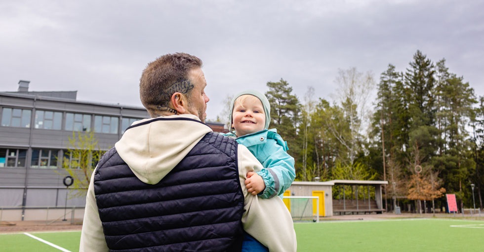 En pappa bakifrån med ett barn på armen som tittar in i kameran. Bilden är tagen på hösten på en fotbollsplan framför en skola.