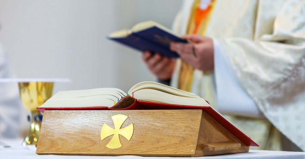 Närbild på en bibel och en nattvardskalk. I bakgrunden står en präst i prästkläder med en bok i handen.