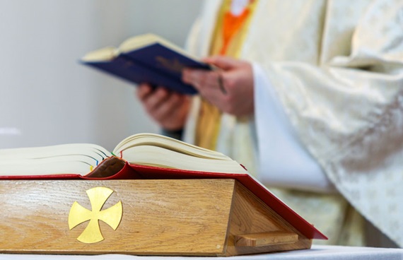 Närbild på en bibel och en nattvardskalk. I bakgrunden står en präst i prästkläder med en bok i handen.