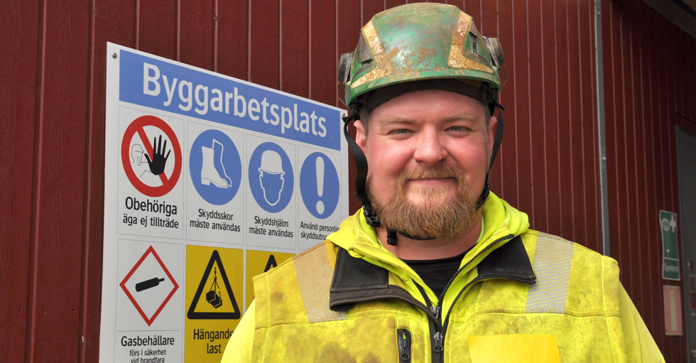 Anton Nijohsson i skyddkläder och skyddshjälm framför en informationsskylt för byggarbetsplats.