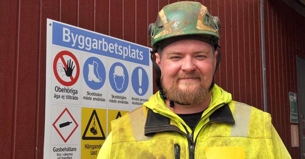Anton Nijohsson i skyddkläder och skyddshjälm framför en informationsskylt för byggarbetsplats.