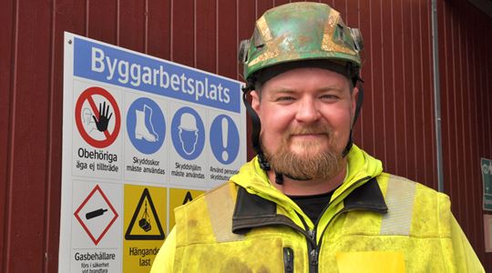 Anton Nijohsson i skyddkläder och skyddshjälm framför en informationsskylt för byggarbetsplats.