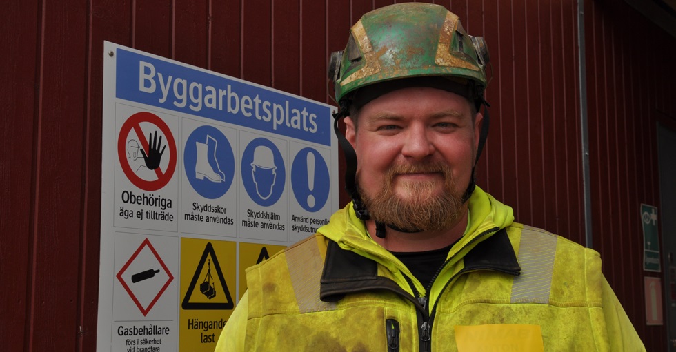 Anton Nijohsson i skyddkläder och skyddshjälm framför en informationsskylt för byggarbetsplats.
