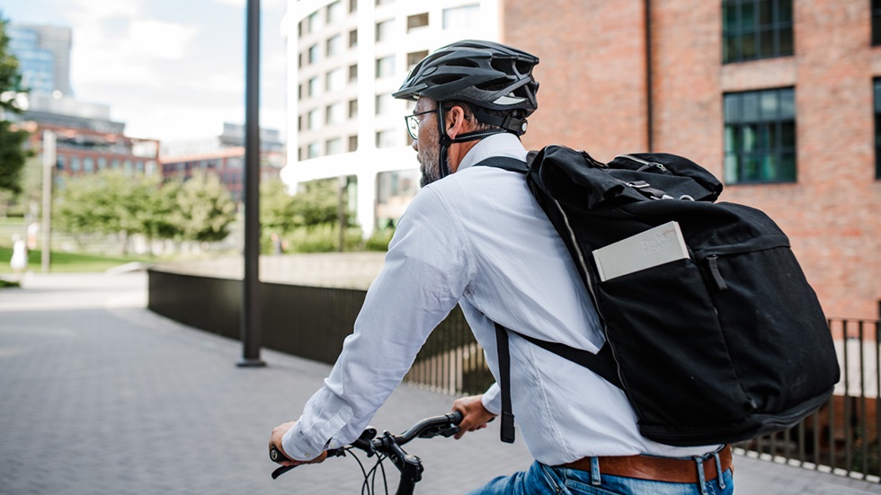 Man i ljusblå skjorta, ryggsäck och cykelhjälm som cyklar i stadsmiljö. 