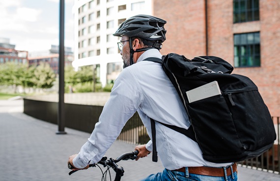 Man i ljusblå skjorta, ryggsäck och cykelhjälm som cyklar i stadsmiljö.