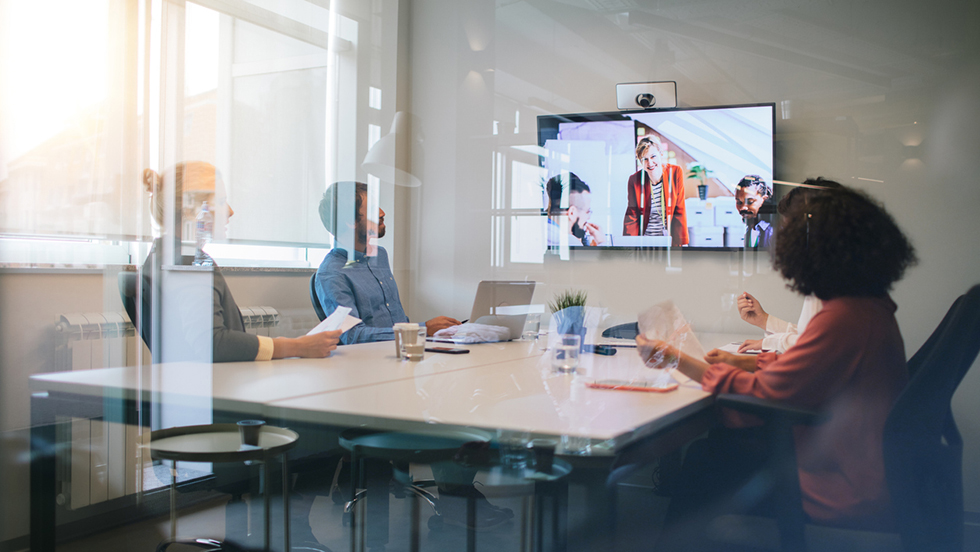 Tre kollegor sitter runt ett mötesbord och har ett digitalt möte med tre andra kollegor på en skärm.