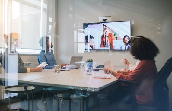 Tre kollegor sitter runt ett mötesbord och har ett digitalt möte med tre andra kollegor på en skärm.