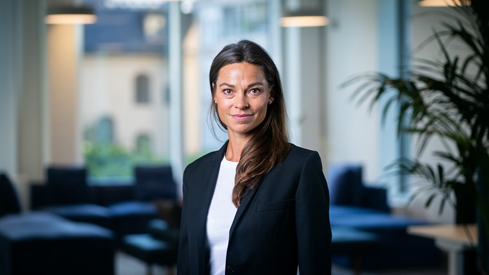 Mörkhårig kvinna i kontorslandskap som tittar in i kameran och ler trevligt.