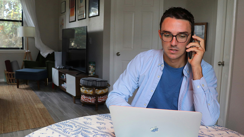 En man sitter vid sitt köksbord och pratar i mobilen med sin laptop framför sig.
