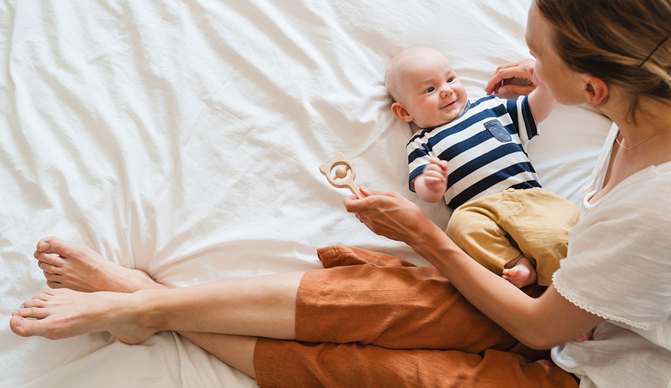 Glad mamma som leker med bebis på en vit säng.