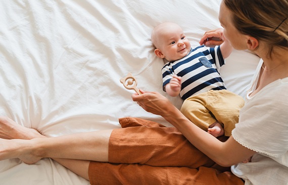 Glad mamma som leker med bebis på en vit säng.