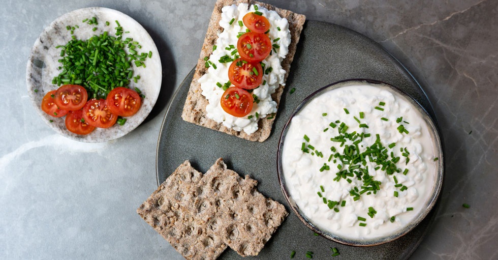 Bild fotad ovanifrån på ett bord med knäckebröd med keso, tomater och färska örter.