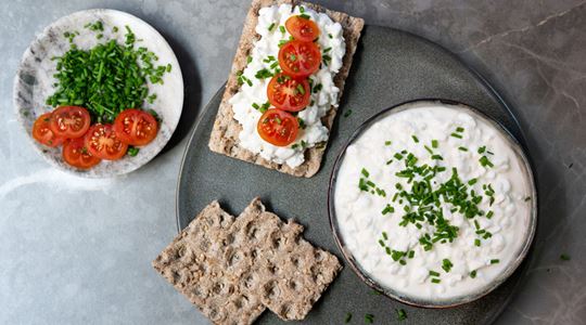 Bild fotad ovanifrån på ett bord med knäckebröd med keso, tomater och färska örter.
