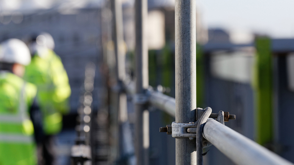 Byggnadsställningar och i bakgrunden syns två byggarbetare med varselväst och hjälmar.
