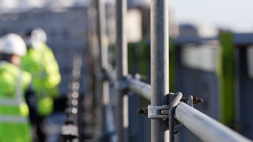 Byggnadsställningar och i bakgrunden syns två byggarbetare med varselväst och hjälmar.
