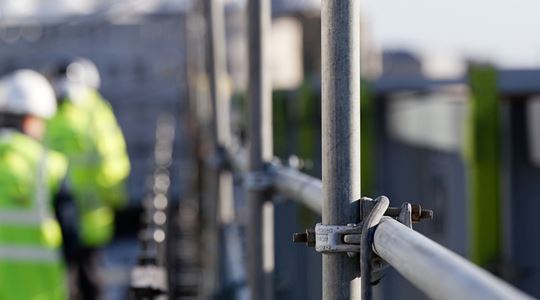 Byggnadsställningar och i bakgrunden syns två byggarbetare med varselväst och hjälmar.