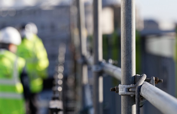 Byggnadsställningar och i bakgrunden syns två byggarbetare med varselväst och hjälmar.