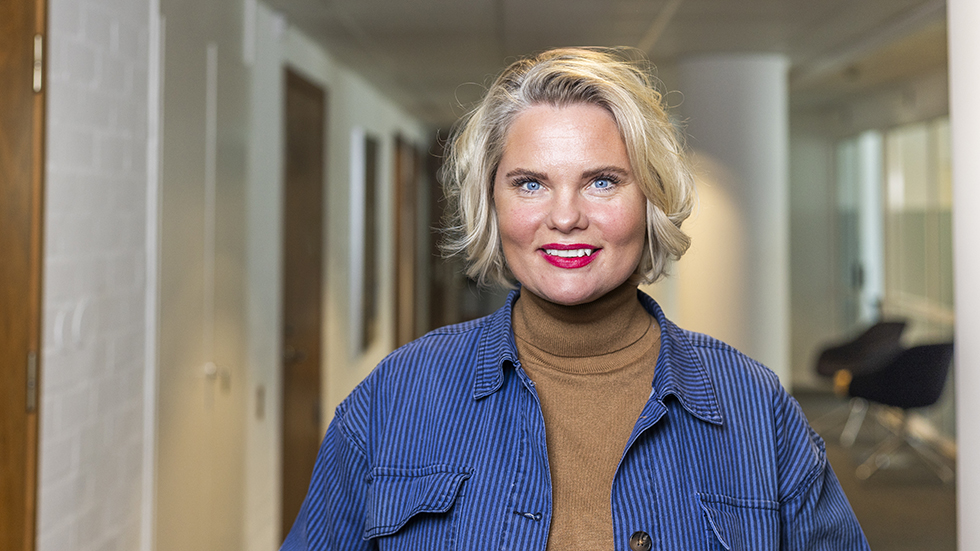 Ljushårig kvinna i korridor på ett kontor ler brett mot kameran.