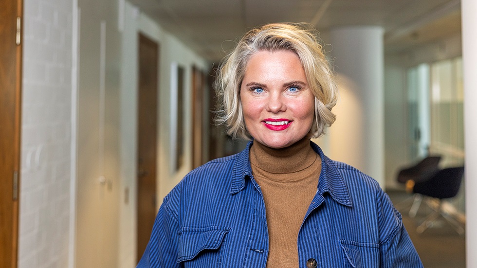 Ljushårig kvinna i korridor på ett kontor ler brett mot kameran.