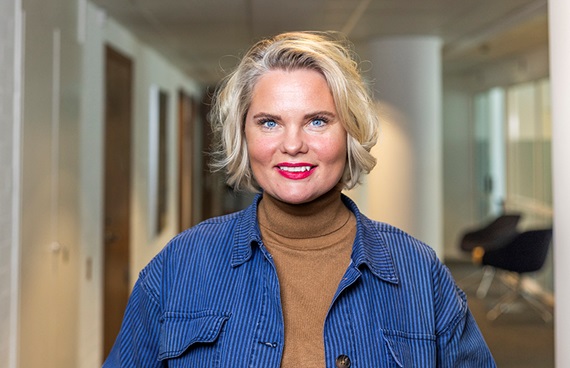 Ljushårig kvinna i korridor på ett kontor ler brett mot kameran.