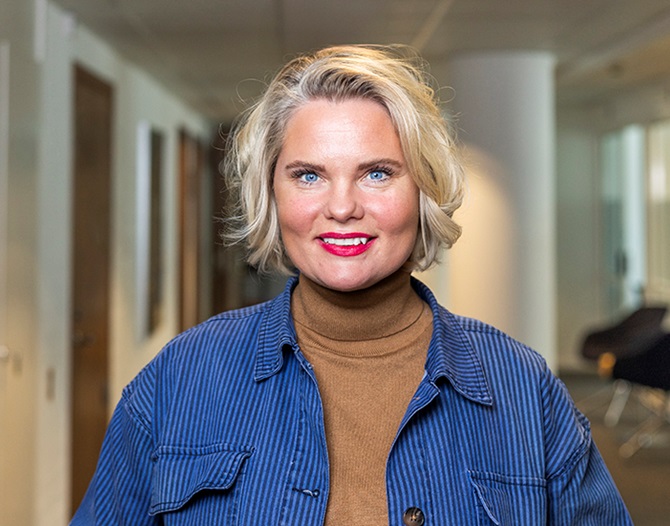 Ljushårig kvinna i korridor på ett kontor ler brett mot kameran.