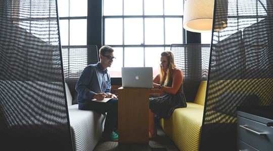 Två kollegor sitter mittemot varandra och tittar på en skärm som står på ett litet bord mellan dem.