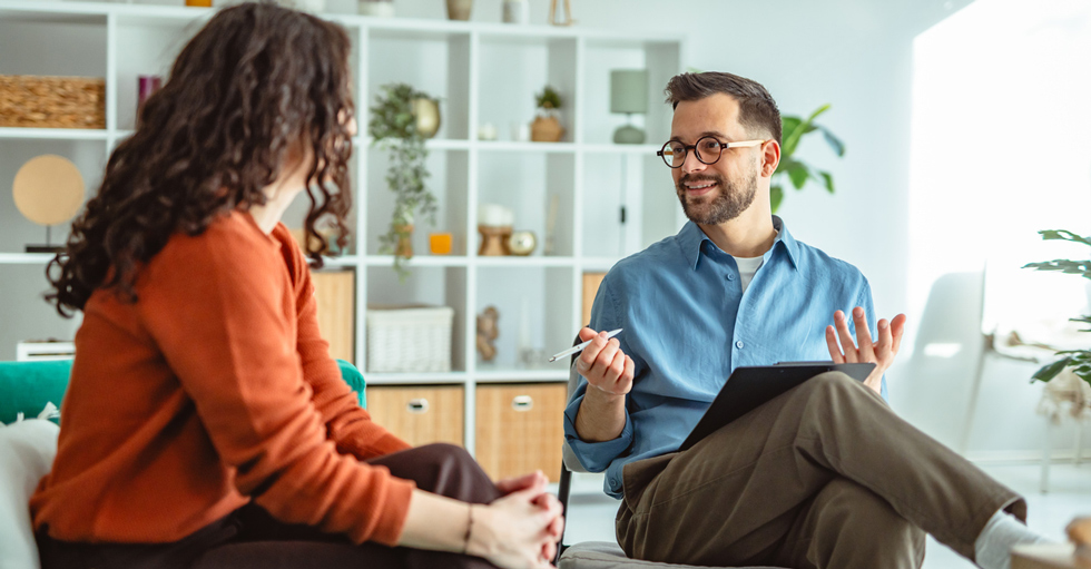 En kvinna i roströd kofta med ryggen mot kameran och en man med blå skjorta och glasögon samtalar i ett ljust rum med en vit bokhylla i bakgrunden.