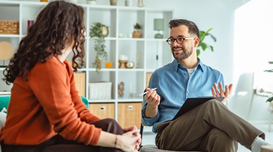 En kvinna i roströd kofta med ryggen mot kameran och en man med blå skjorta och glasögon samtalar i ett ljust rum med en vit bokhylla i bakgrunden.