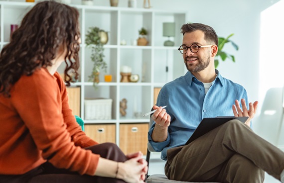 En kvinna i roströd kofta med ryggen mot kameran och en man med blå skjorta och glasögon samtalar i ett ljust rum med en vit bokhylla i bakgrunden.