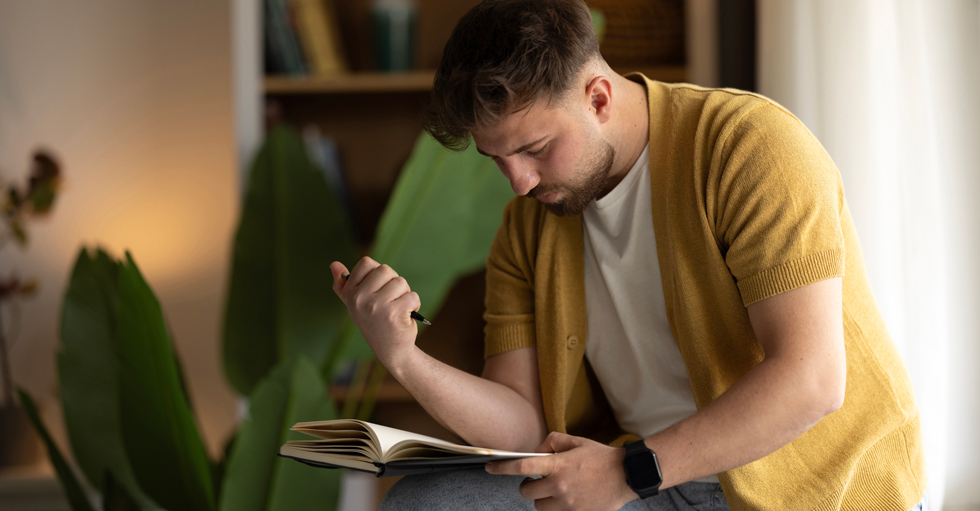 En man sitter böjd över en anteckningsbok med en penna i handen.