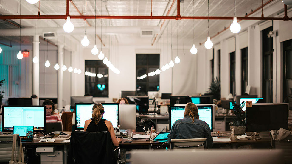 Människor sitter på rad och arbetar framför skärmar i ett stort kontorslandskap.