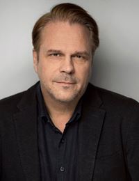 Porträtt av Eric Donell i svart skjorta och kavaj mot en grå bakgrund.