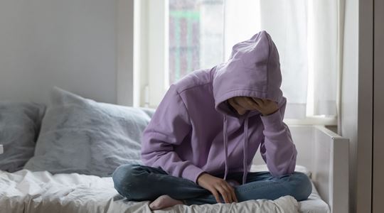 En ung person, klädd i lila huvtröja, sitter framåtböjd på en säng och lutar huvudet i handen.
