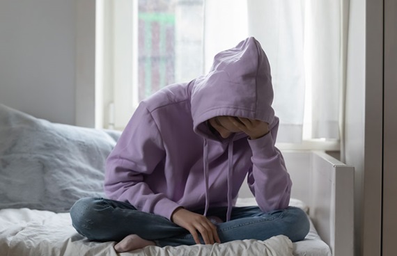En ung person, klädd i lila huvtröja, sitter framåtböjd på en säng och lutar huvudet i handen.