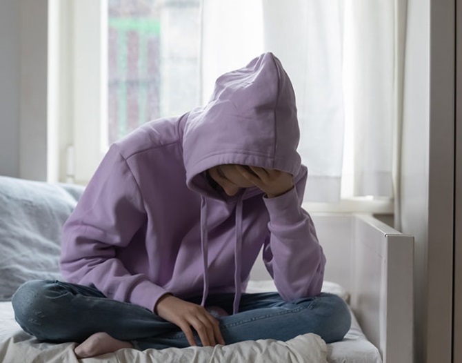 En ung person, klädd i lila huvtröja, sitter framåtböjd på en säng och lutar huvudet i handen.