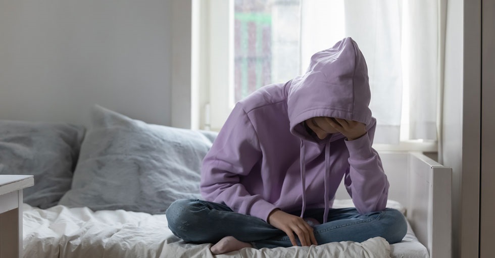 En ung person, klädd i lila huvtröja, sitter framåtböjd på en säng och lutar huvudet i handen.